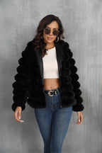 Woman wearing a black faux fur hooded jacket over a white crop top and blue jeans, with sunglasses on.