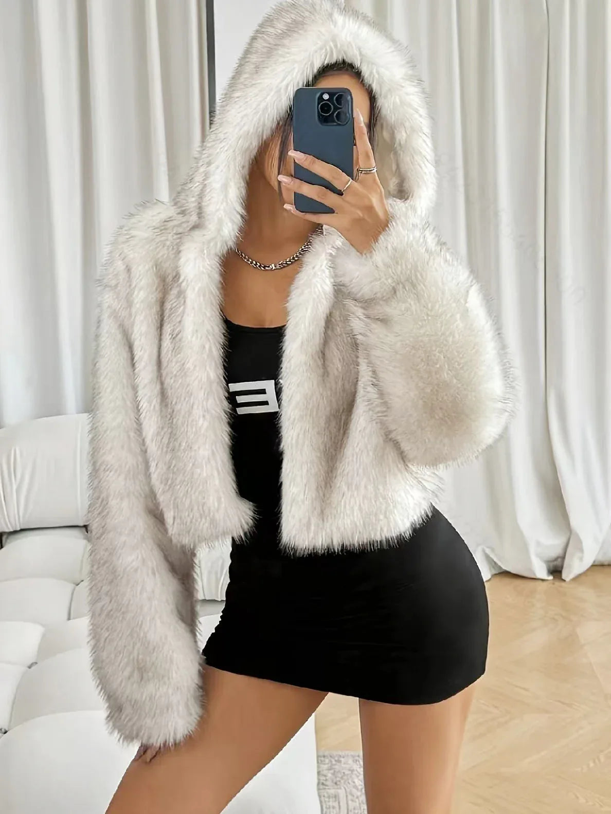 Woman wearing a white faux fur cropped hooded jacket over a black dress, taking a selfie.