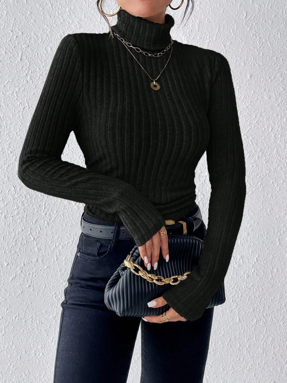 Close-up of a woman wearing a black ribbed turtleneck sweater and black jeans, holding a black clutch with a gold chain.