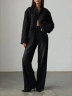 Full body shot of a woman in a black sparkly tweed cropped jacket, black bra, and black wide-leg jeans, standing against a white wall.