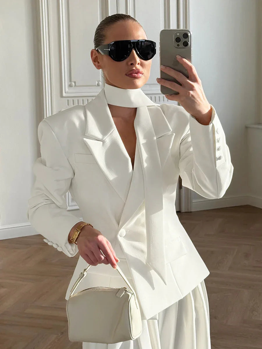 Woman taking a selfie in a white blazer and trousers, holding a small white handbag and wearing sunglasses.