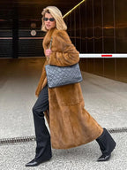 Woman walking in a long brown faux fur coat, black pants, and carrying a quilted black handbag.