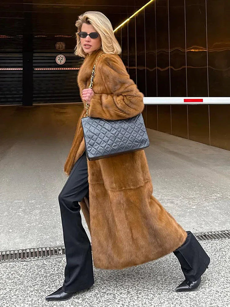 Woman walking in a long brown faux fur coat, black pants, and carrying a quilted black handbag.