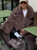 A woman is seated at an outdoor cafe, draped in a plush brown faux fur coat, with her hands resting on the table.