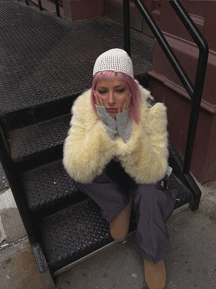 Person with pink hair sitting on stairs, wearing a cream faux fur jacket and grey trousers.