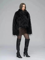 Side view of a woman in a black faux fur jacket, brown wrap dress, sheer black tights, and black knee-high boots.