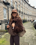 Woman wearing a brown faux fur cropped jacket, white crop top, and brown leggings, smiling at the camera.