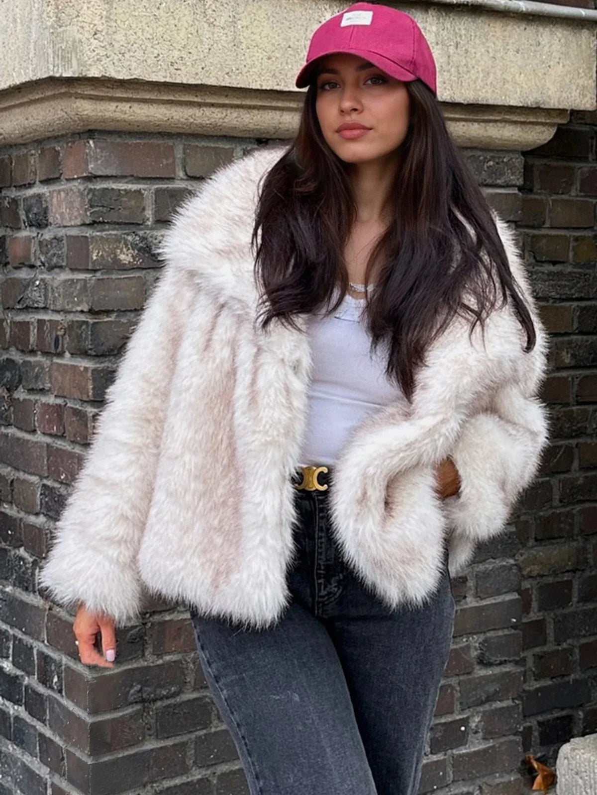 Woman in a cream faux fur jacket and dark jeans, standing against a brick wall.