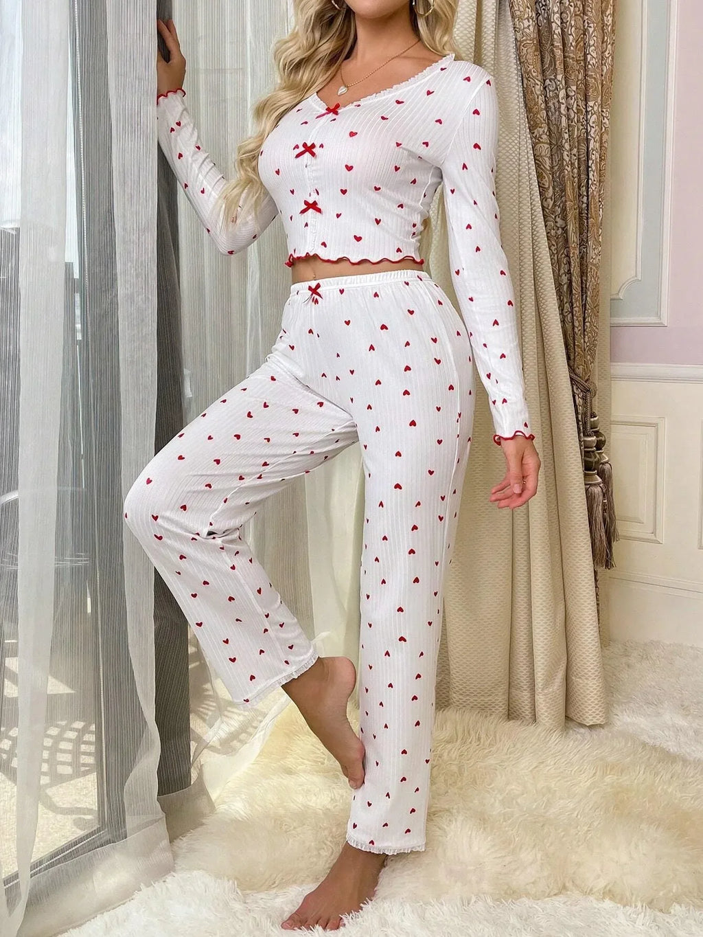 Woman wearing a white long-sleeve cropped pajama top and matching pants with a red heart print, leaning against a window frame.