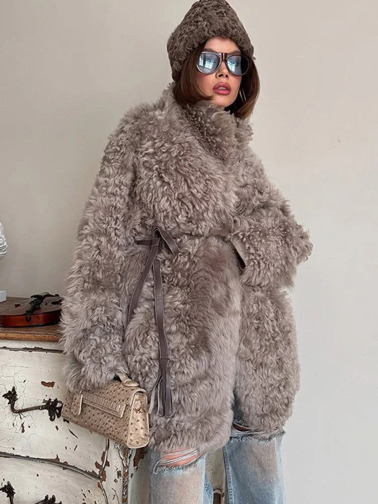 Woman wearing a gray curly faux shearling coat cinched with a belt, brown beanie, and ripped blue jeans, holding a beige clutch bag.