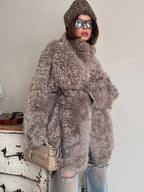 Woman wearing a gray curly faux shearling coat cinched with a belt, brown beanie, and ripped blue jeans, holding a beige clutch bag.