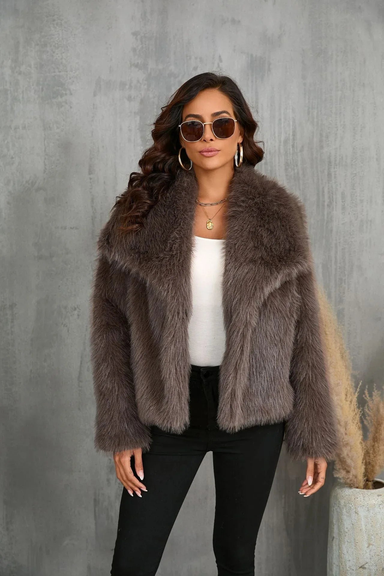 Woman wearing a brown faux fur jacket, white top, and black pants, holding sunglasses.