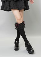Legs of a person wearing black knee-high socks with decorative ruffled lace and rose accents.