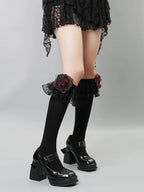 A person wearing black knee-high socks with ruffled black lace trim and a red rose embellishment on each sock, paired with black platform mary jane shoes.