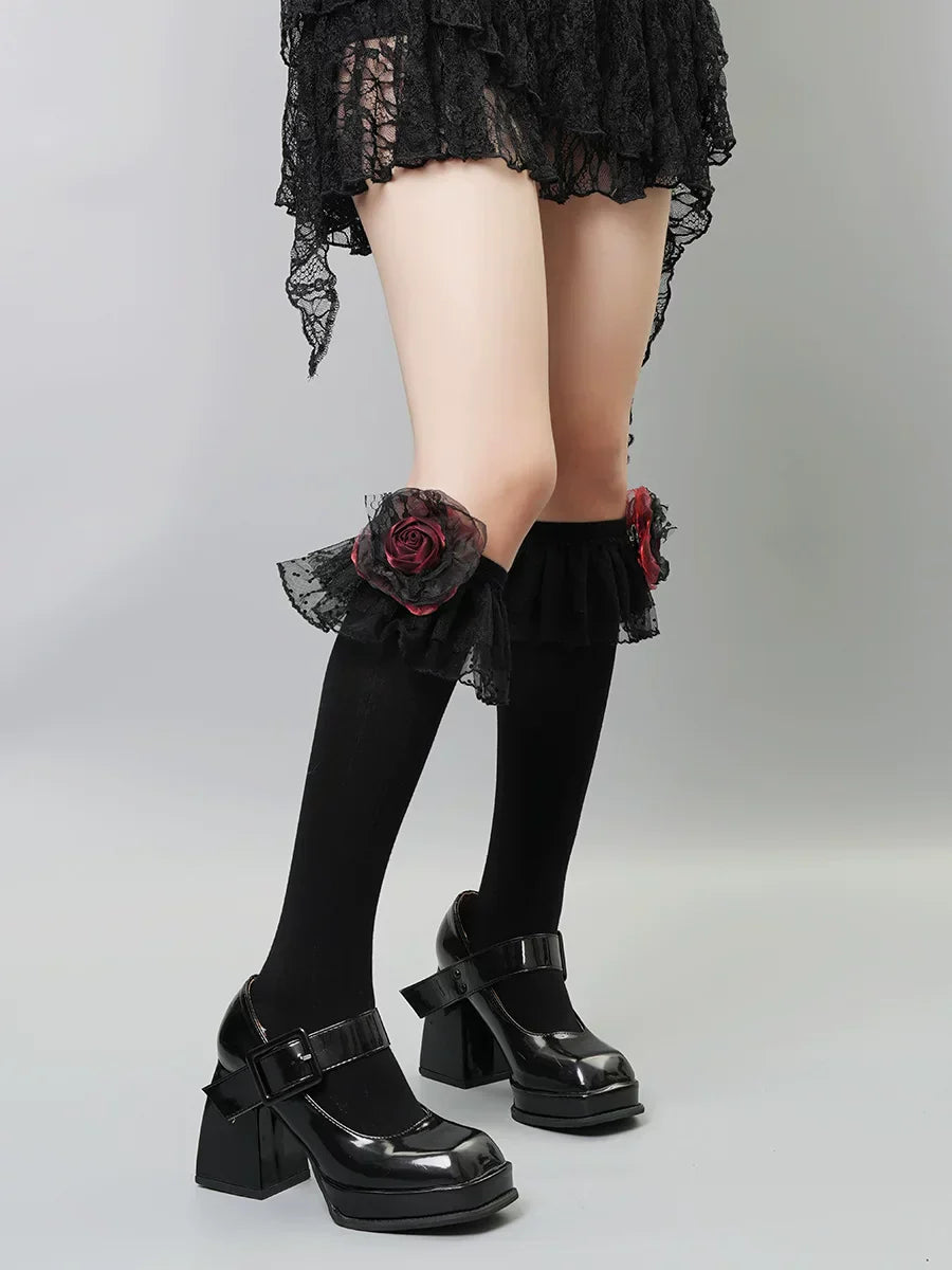 A person wearing black knee-high socks with ruffled black lace trim and a red rose embellishment on each sock, paired with black platform mary jane shoes.