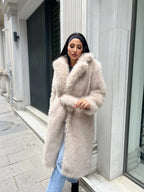 Woman wearing a long, cream-colored faux fur coat with a belted waist and blue jeans, posing outdoors.