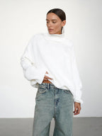 Close-up of a woman wearing an oversized white turtleneck sweater, lifting the hem to show her jeans.