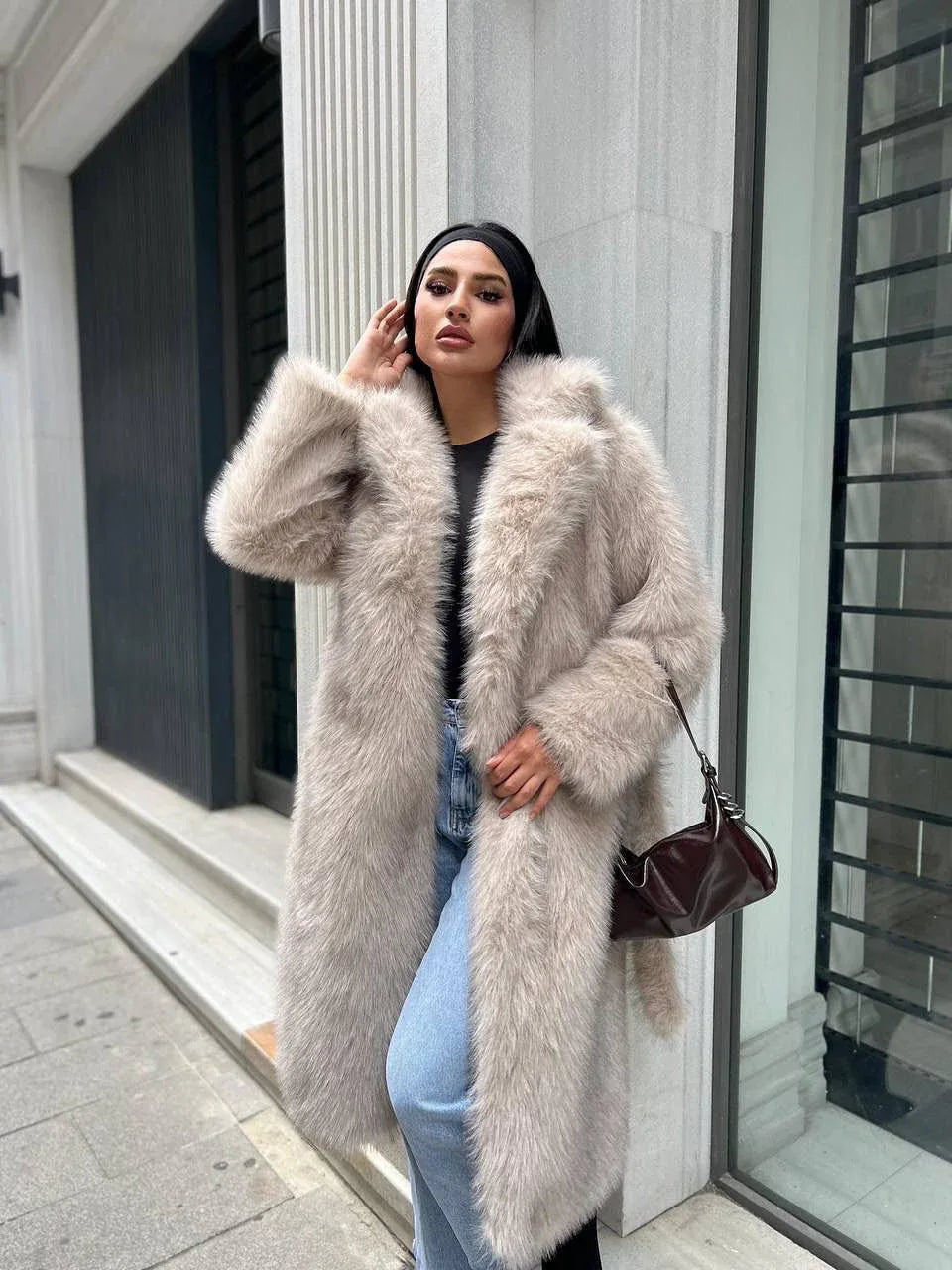 Woman in a long, cream-colored faux fur coat, blue jeans, and a black headband, holding a brown handbag.