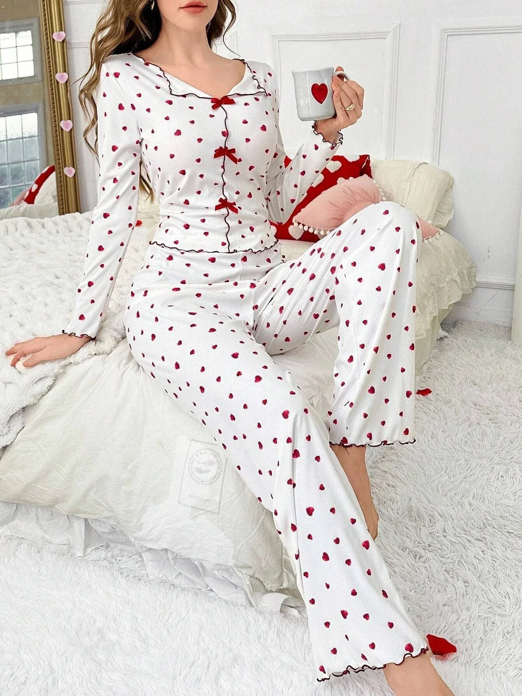 Woman wearing white pajamas with red heart print, sitting on a bed and holding a mug.