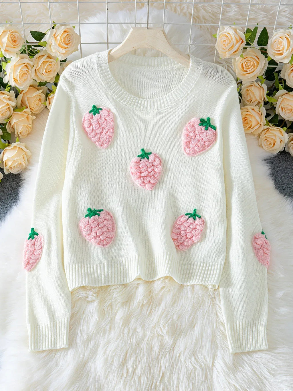 White cropped knit sweater with multiple pink strawberry appliques, displayed on a hanger with a floral background.