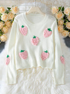 White cropped knit sweater with multiple pink strawberry appliques, displayed on a hanger with a floral background.