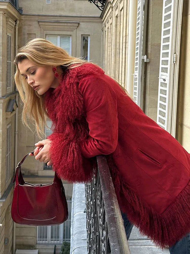 Woman leaning on a balcony railing, wearing a red suede coat with fluffy red fur trim and cuffs, looking down.