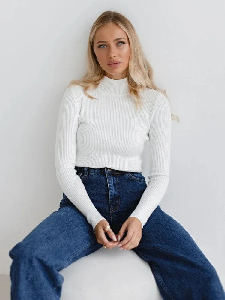 Woman wearing a white ribbed mock neck cropped sweater and dark wash wide-leg jeans, sitting on a white ottoman. Main image