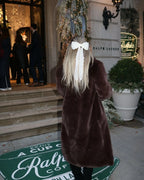 Rear view of a woman wearing a long brown faux fur coat with a large white lace bow in her blonde hair.