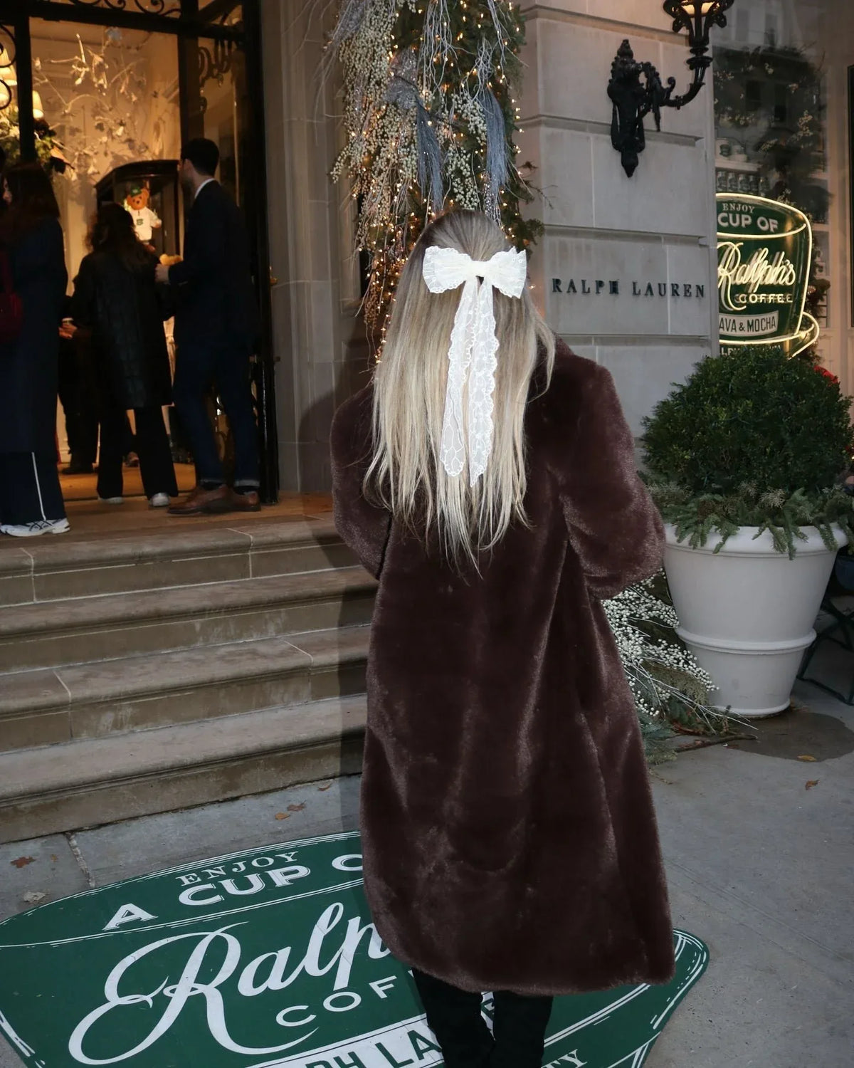 Rear view of a woman wearing a long brown faux fur coat with a large white lace bow in her blonde hair.