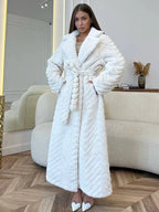 Woman wearing a white faux fur long coat with a chevron pattern and a belted waist, standing indoors in a modern living room.