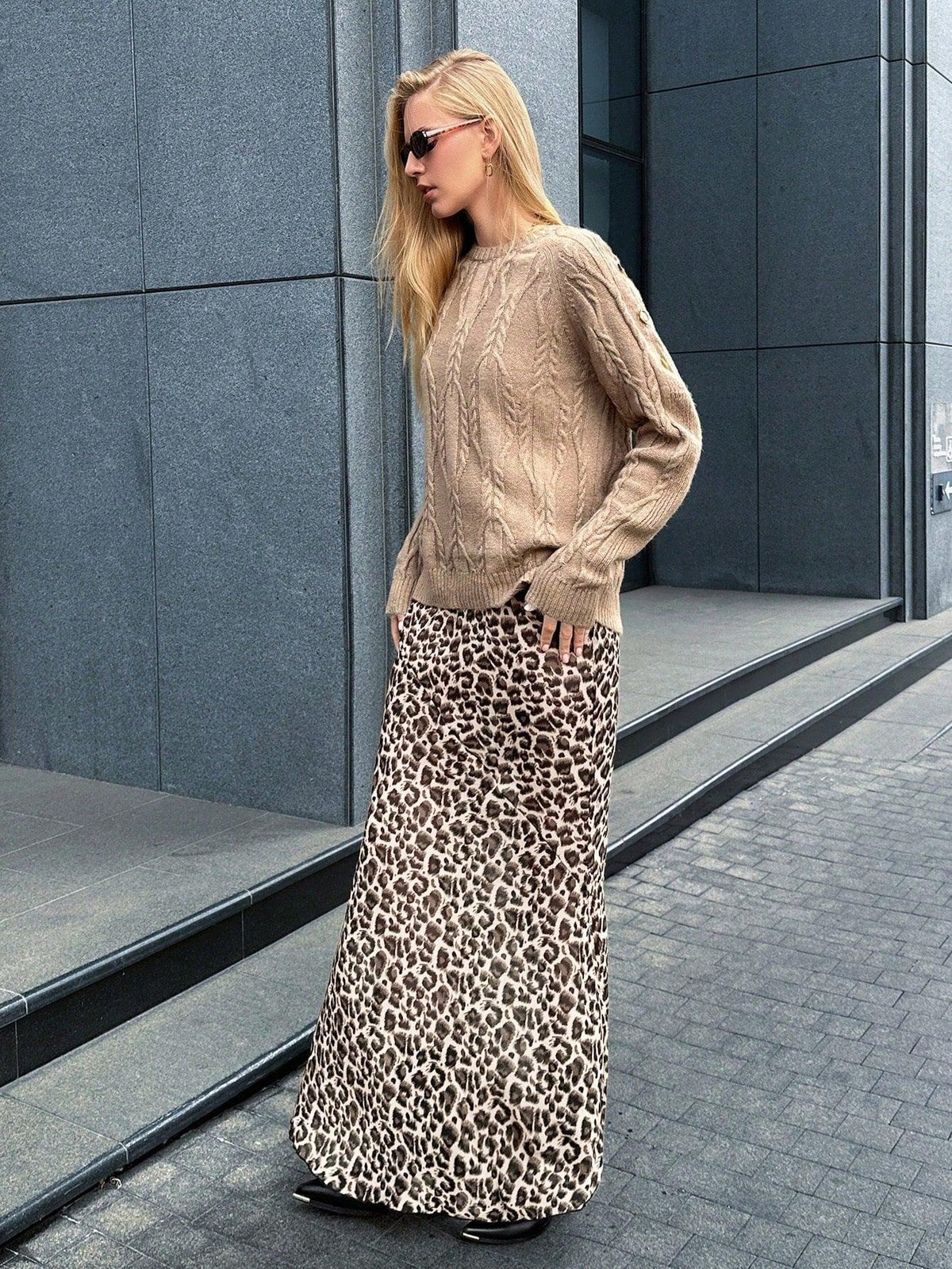 Woman posing in a tan cable knit sweater and leopard print maxi skirt, with hands in pockets.