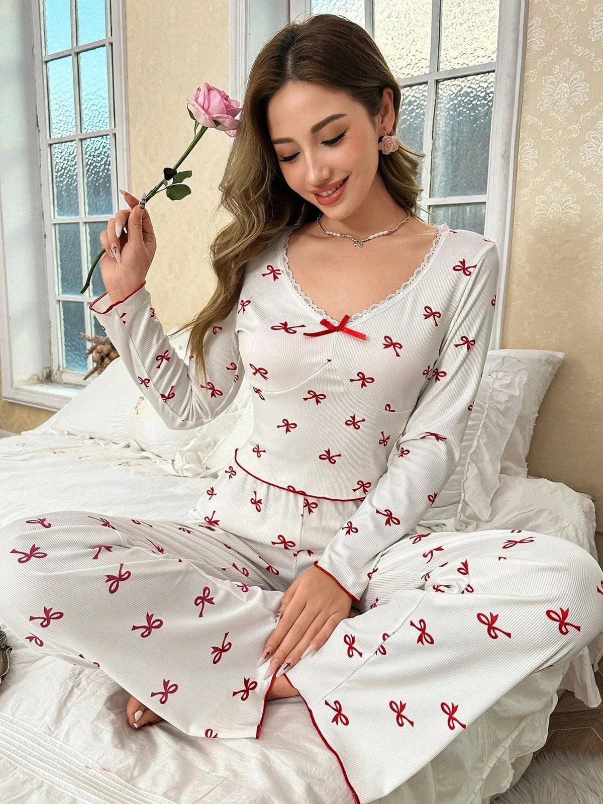 woman wearing white and red bow print pajama set sitting on a bed holding a rose