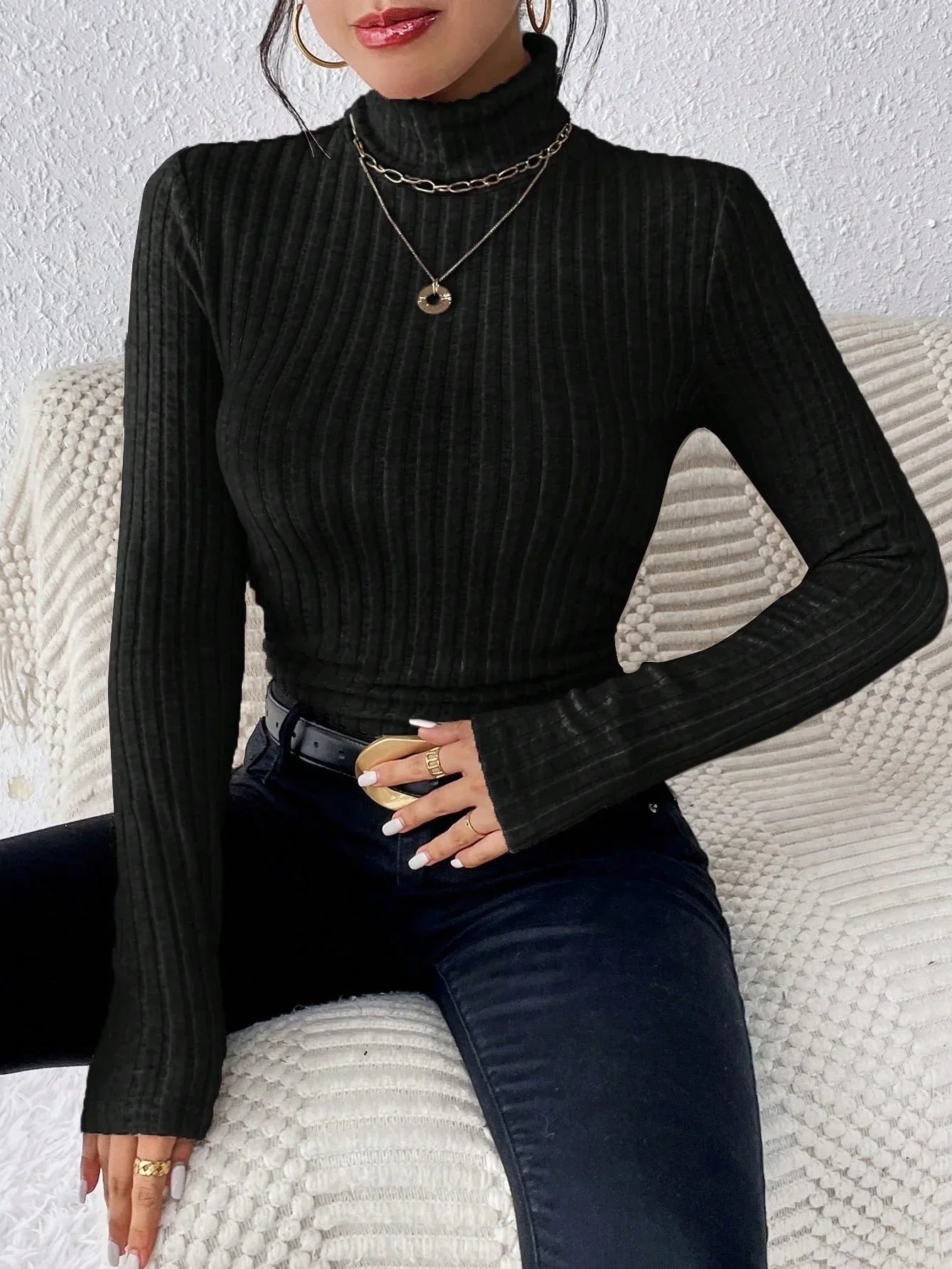 Woman wearing a black ribbed turtleneck sweater and black jeans, accessorized with gold necklaces and rings.