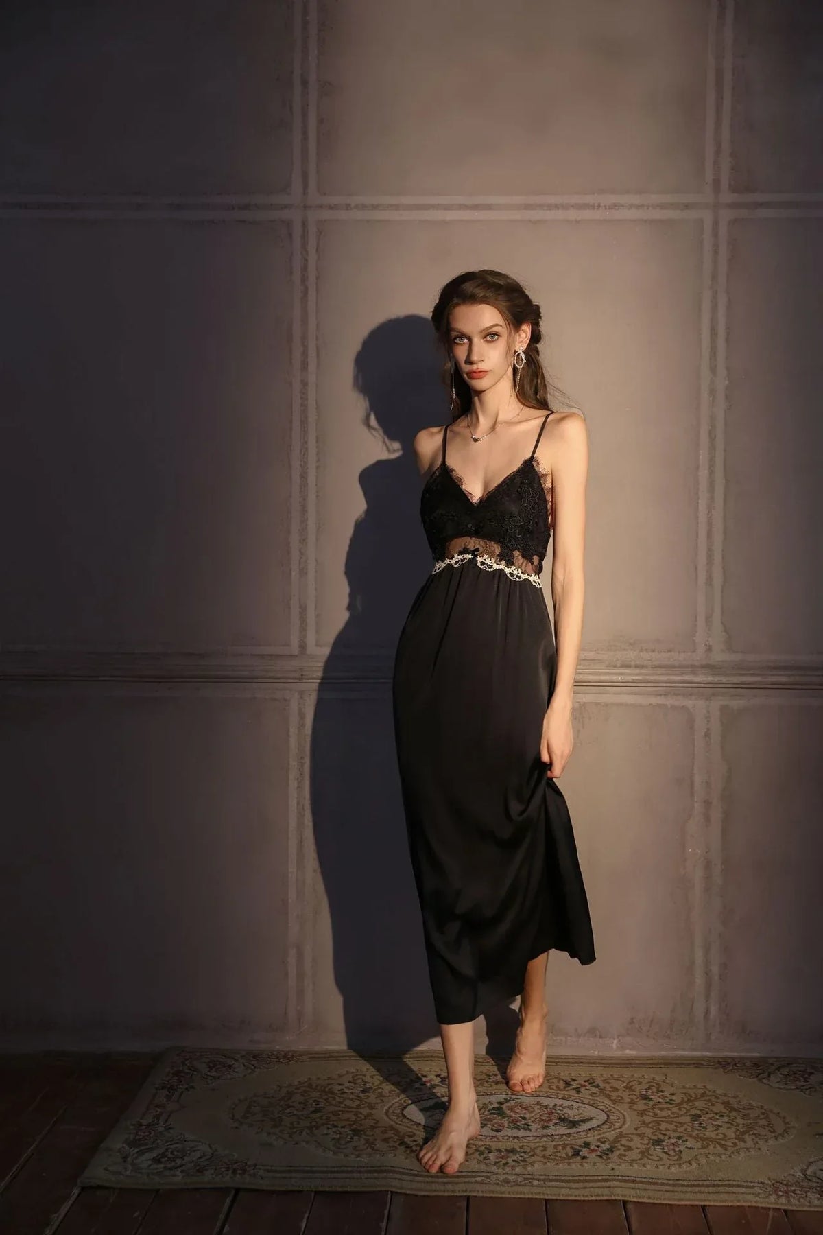 Woman wearing a black satin slip dress with lace trim, holding the hem of the dress and looking at the camera.