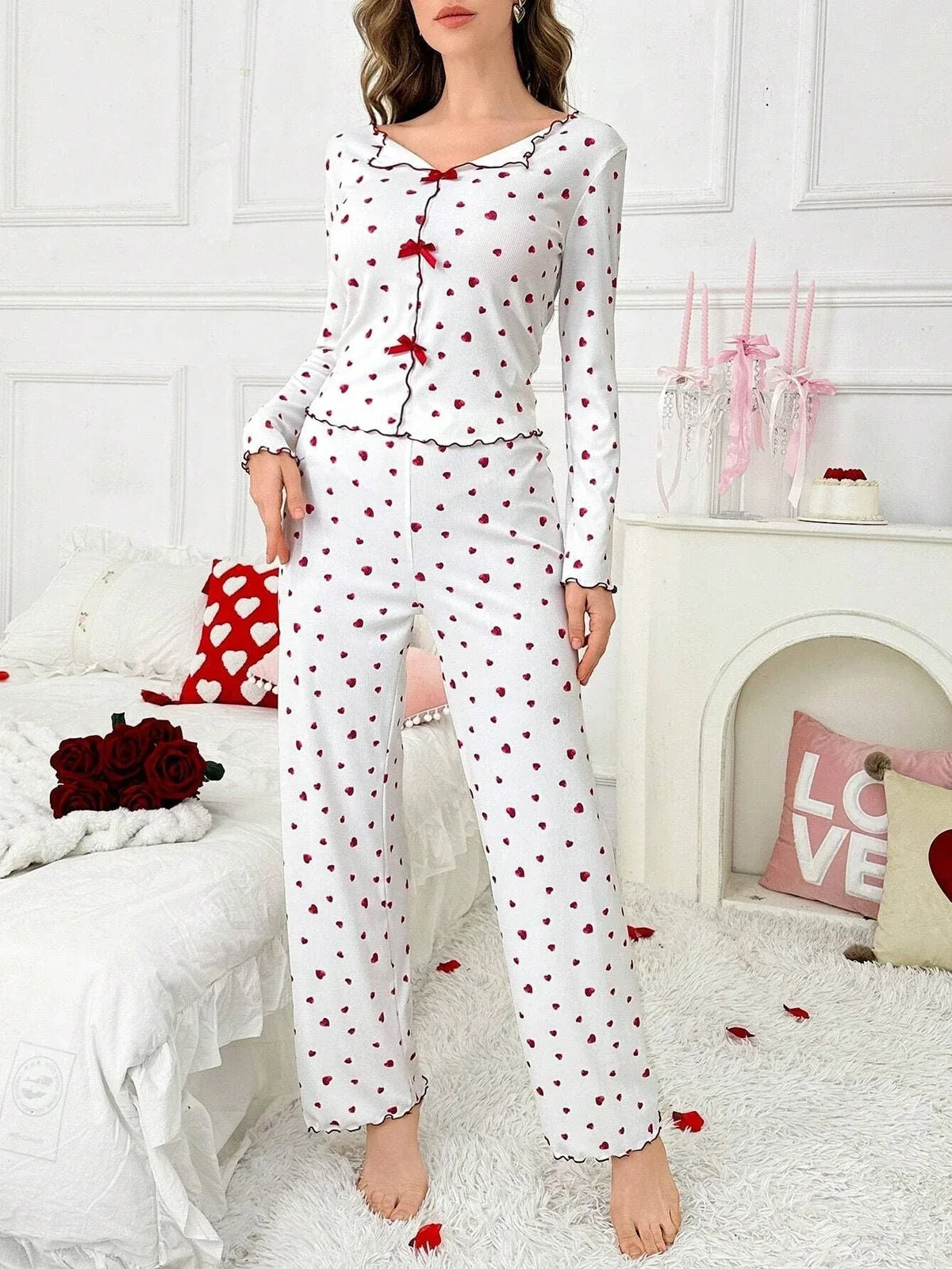 Woman wearing white pajamas with red heart print and red bows, standing in a bedroom. Main image