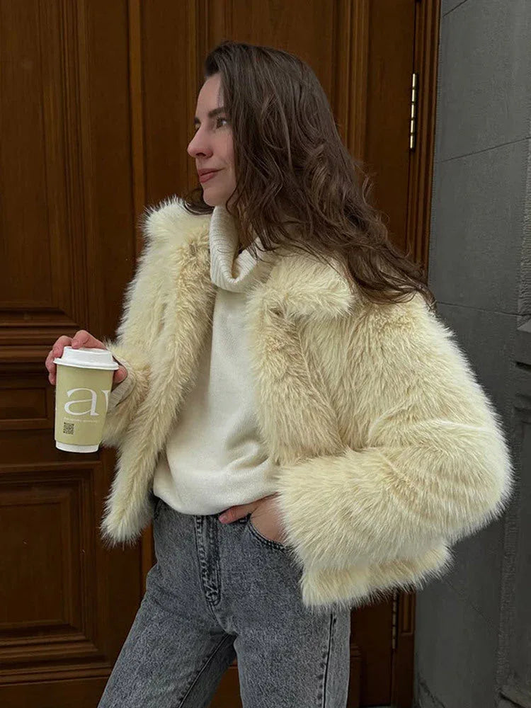 Woman wearing a cream faux fur jacket and grey jeans, holding a coffee cup.