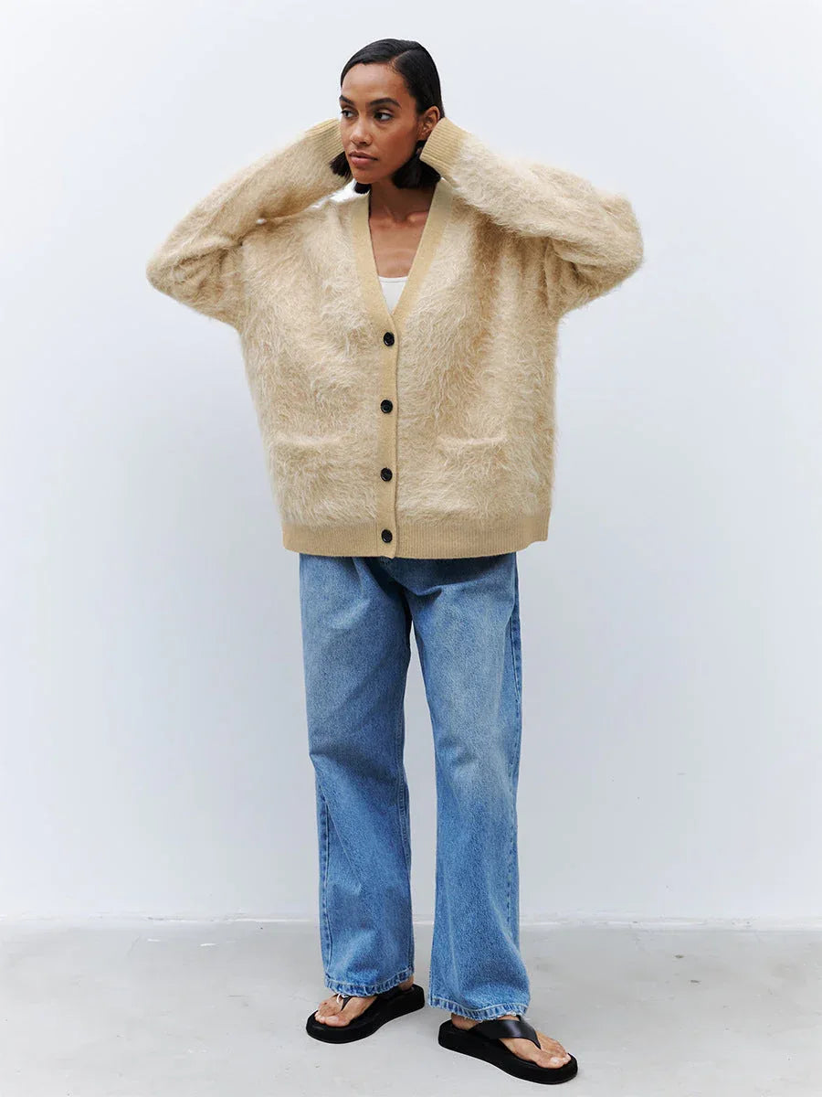 Full body shot of a woman wearing a beige fuzzy V-neck button-up cardigan with two front pockets and blue jeans.