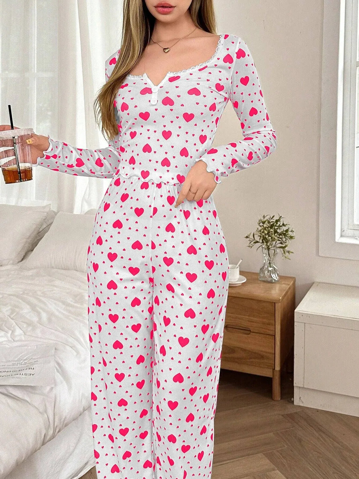 Woman wearing a white long-sleeved pajama top and pants with a pink heart print, holding a drink. Main image
