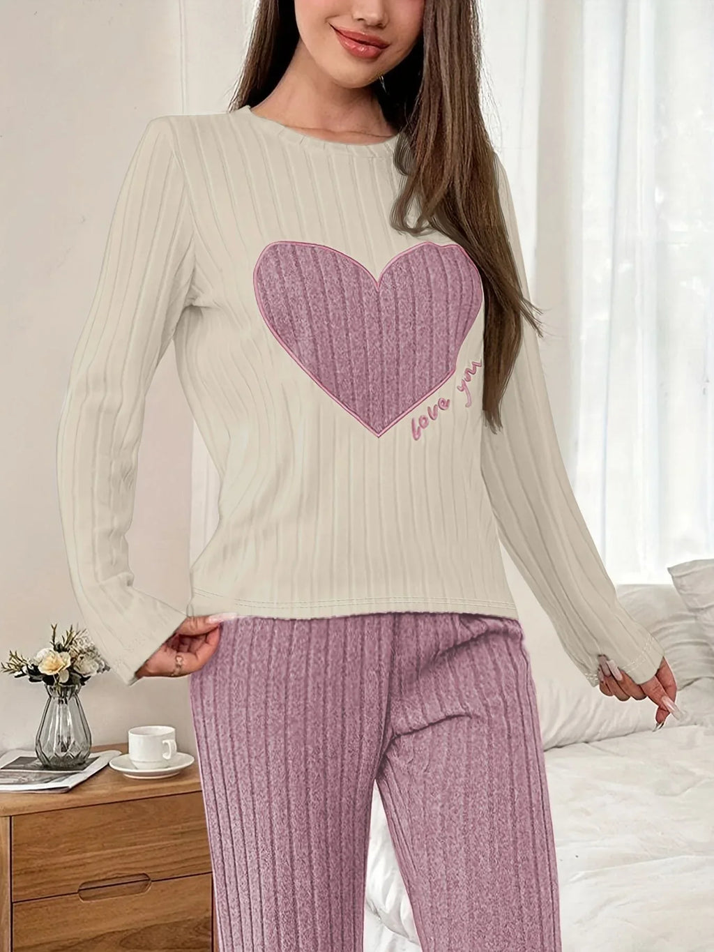 Woman standing in a bedroom, adjusting her pajama pants, wearing a cream long-sleeve pajama top with a pink heart graphic and "love you" text, and matching ribbed purple pajama pants.