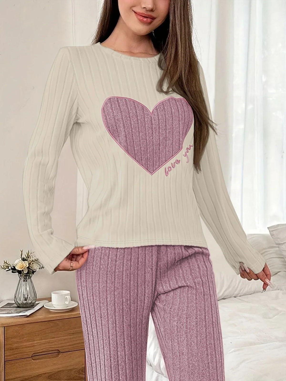 Woman standing in a bedroom, adjusting her pajama pants, wearing a cream long-sleeve pajama top with a pink heart graphic and "love you" text, and matching ribbed purple pajama pants.
