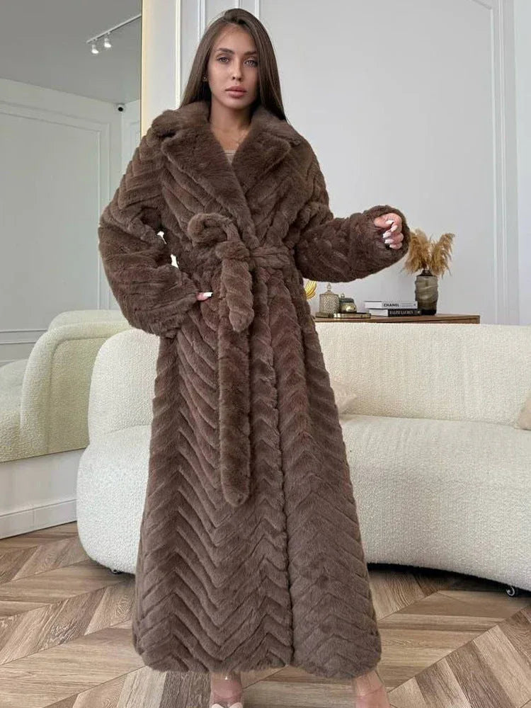 Woman wearing a brown faux fur long coat with a chevron pattern and a belted waist, standing indoors in a modern living room.
