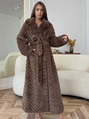 Woman wearing a brown faux fur long coat with a chevron pattern and a belted waist, standing indoors in a modern living room.