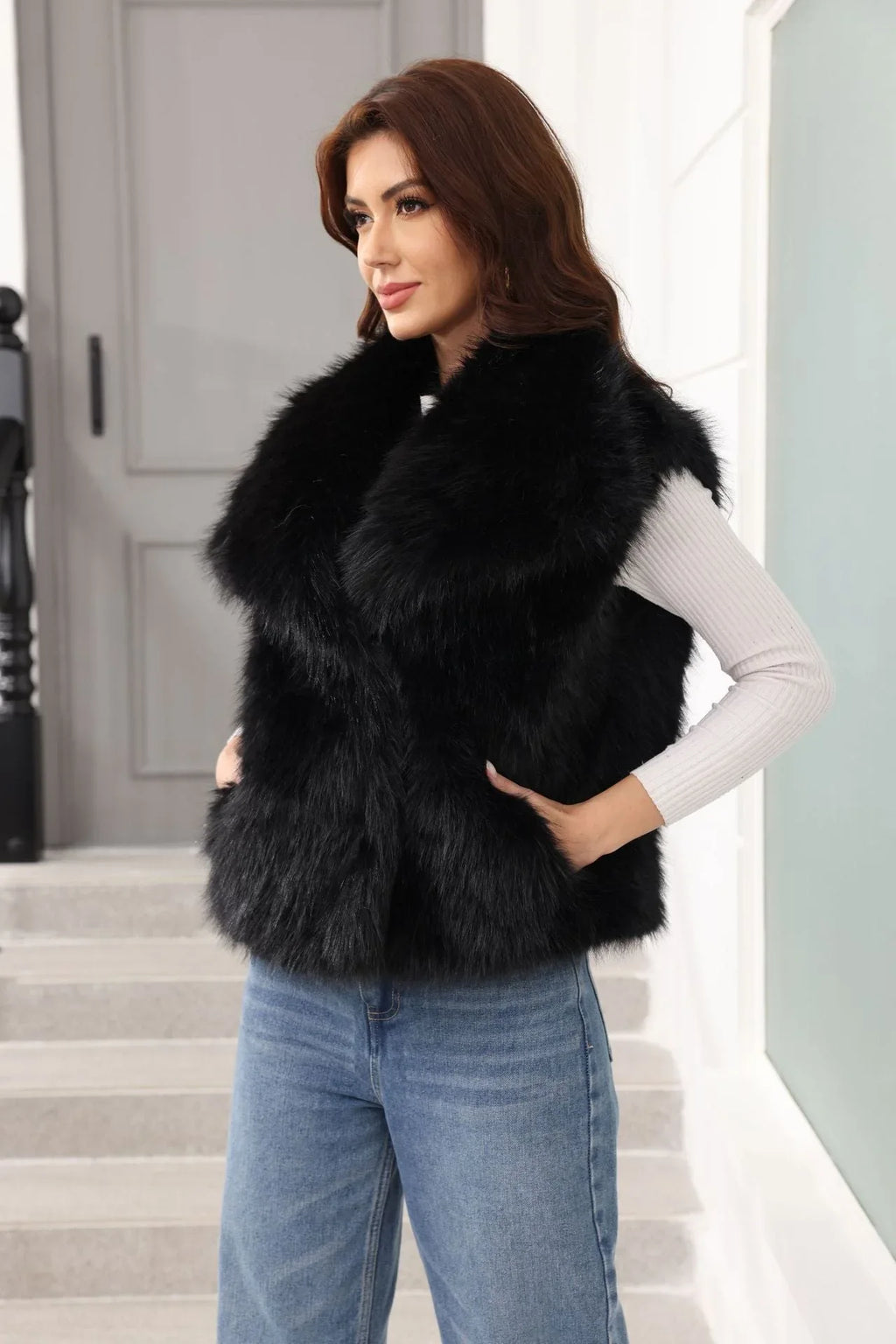 Woman wearing a black faux fur vest over a white long-sleeved shirt and blue jeans, with one hand on hip