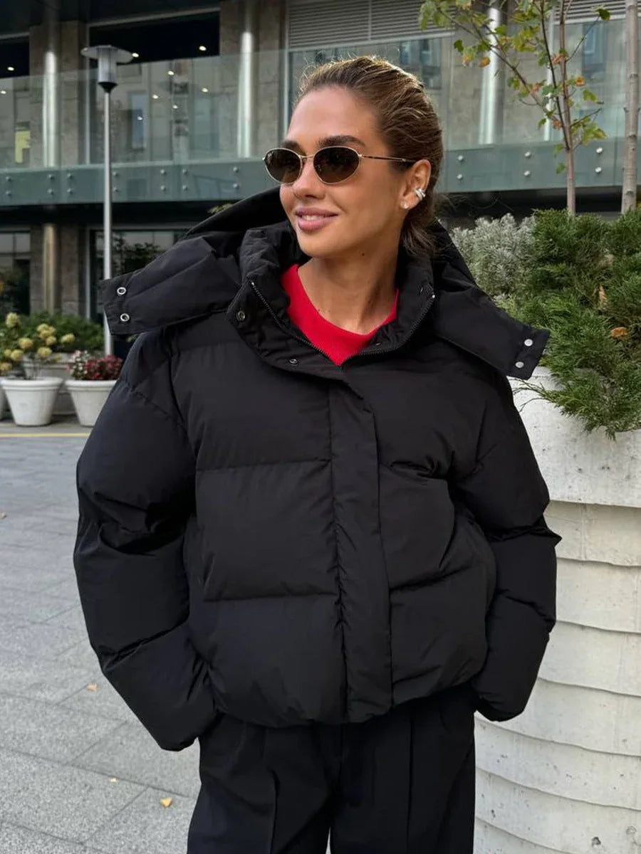 Woman wearing a black cropped puffer jacket with a hood, red t-shirt, and black pants, standing outdoors.