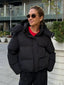 Woman wearing a black cropped puffer jacket with a hood, red t-shirt, and black pants, standing outdoors.