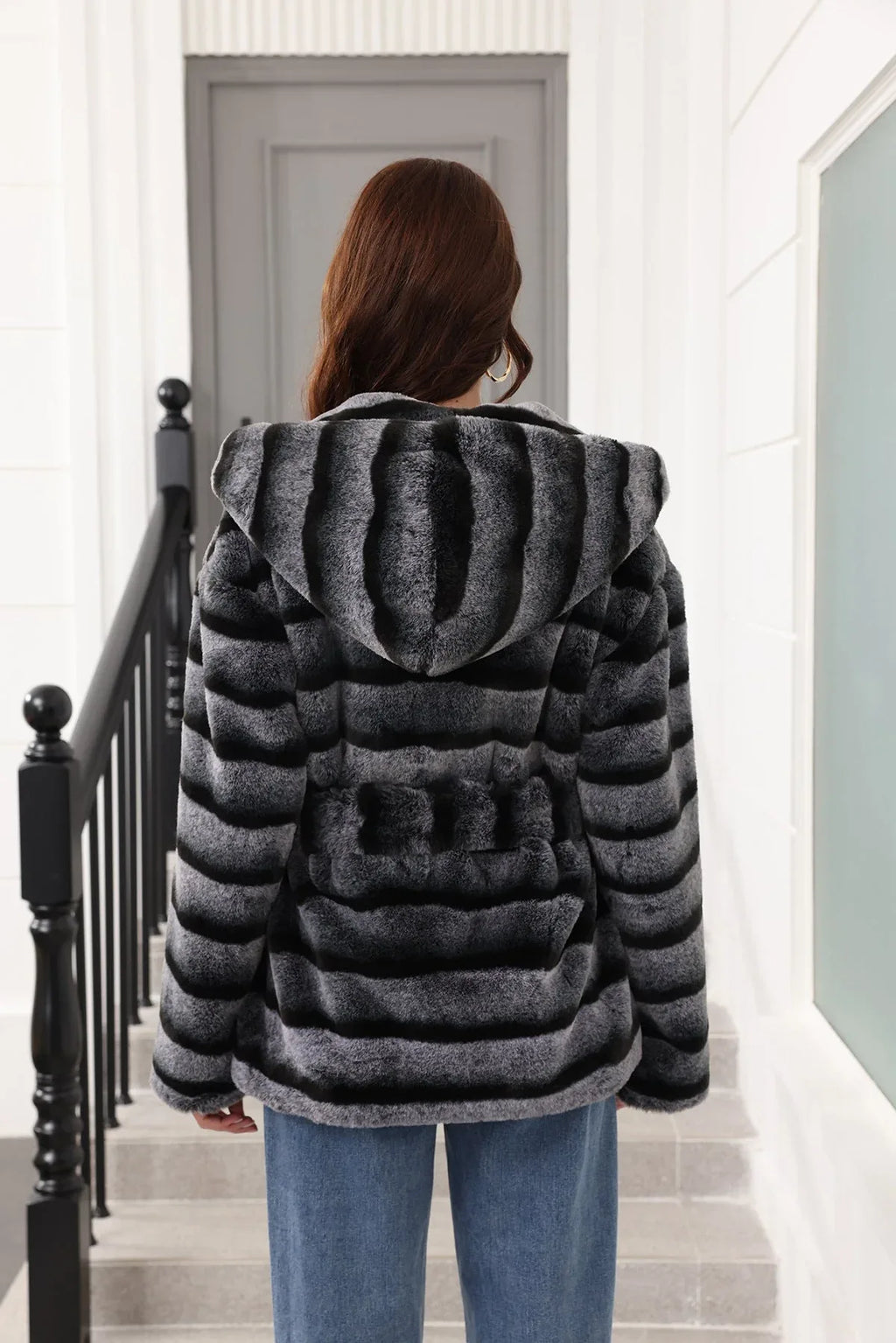 Back view of a woman wearing a hooded faux fur jacket with horizontal black and grey stripes and a belted waist.