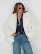 Woman wearing a white faux fur jacket, denim shirt, belt, and blue jeans, with hands in pockets.