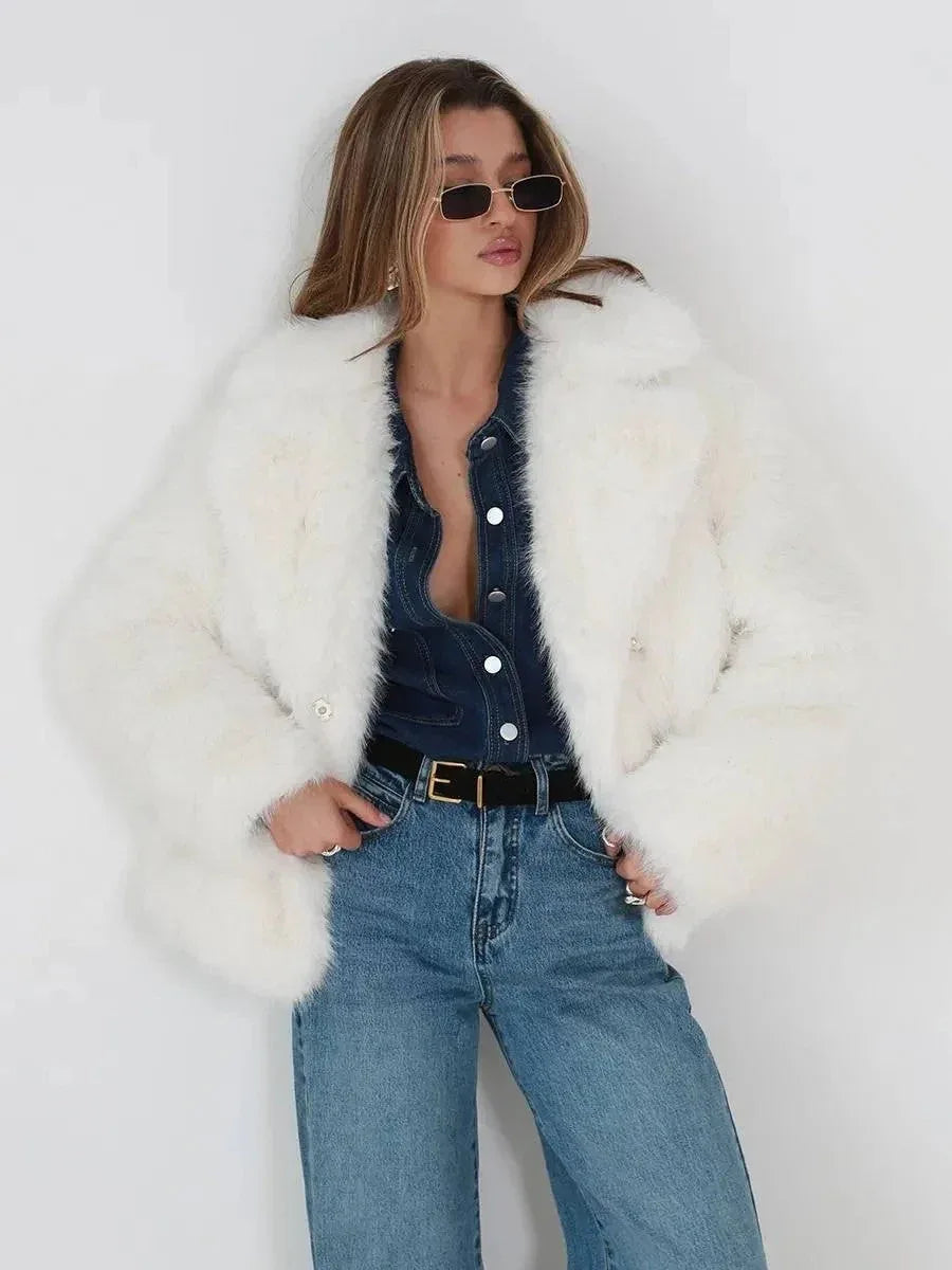 Woman wearing a white faux fur jacket, denim shirt, belt, and blue jeans, with hands in pockets.