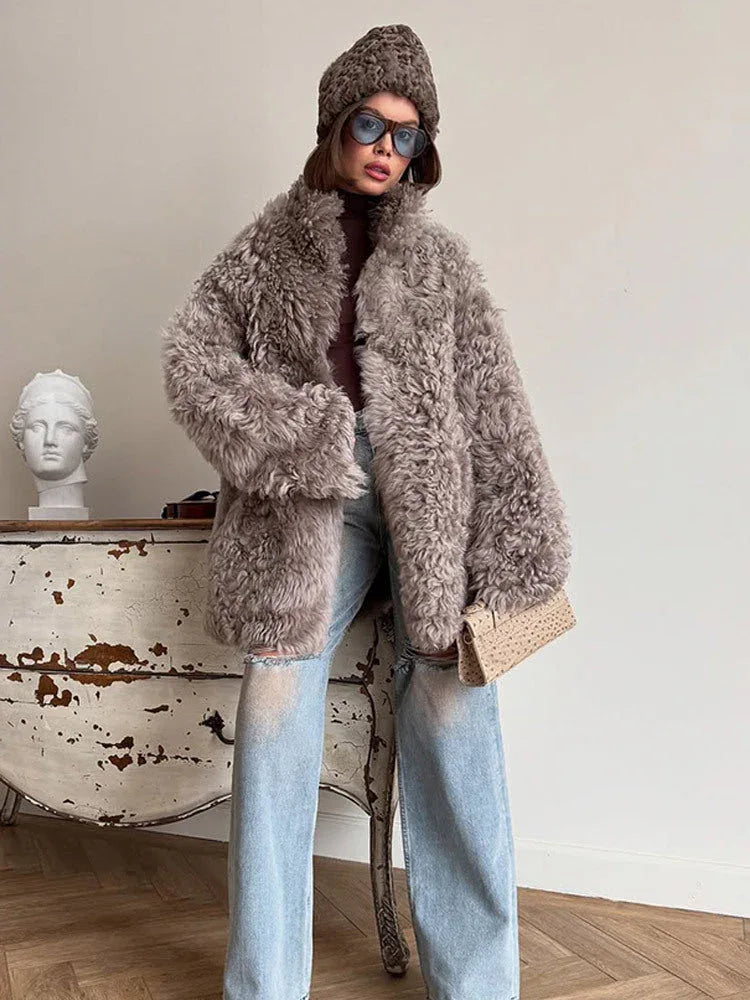 Woman wearing a gray curly faux shearling coat with a brown beanie and ripped blue jeans, holding a beige clutch bag. Main image