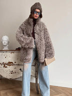 Woman wearing a gray curly faux shearling coat with a brown beanie and ripped blue jeans, holding a beige clutch bag.